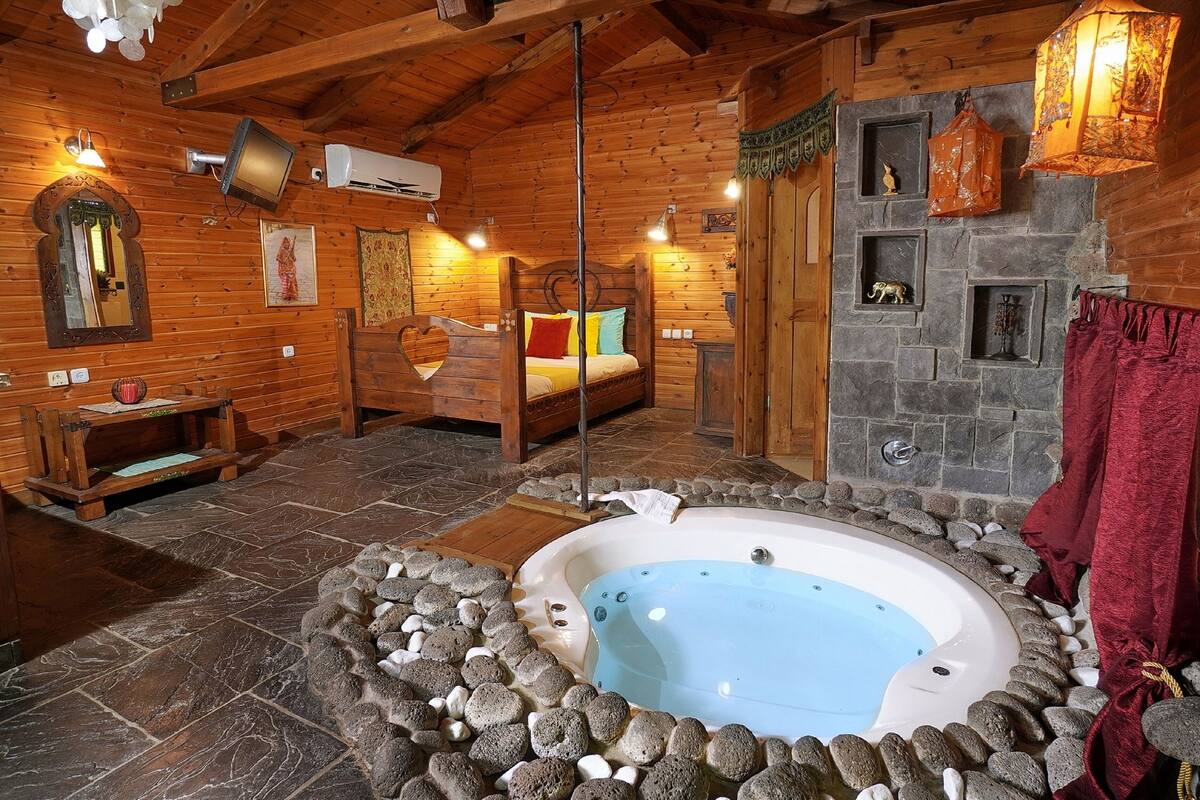 A log cabin bedroom with a stone-tiled floor, a wooden bed, a stone hot tub, and a stone fireplace.