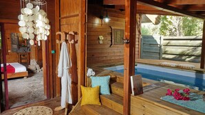 Una cabaña de madera con una cama, un banco de madera con cojines, una mesita con un libro y flores, y vistas a una piscina.