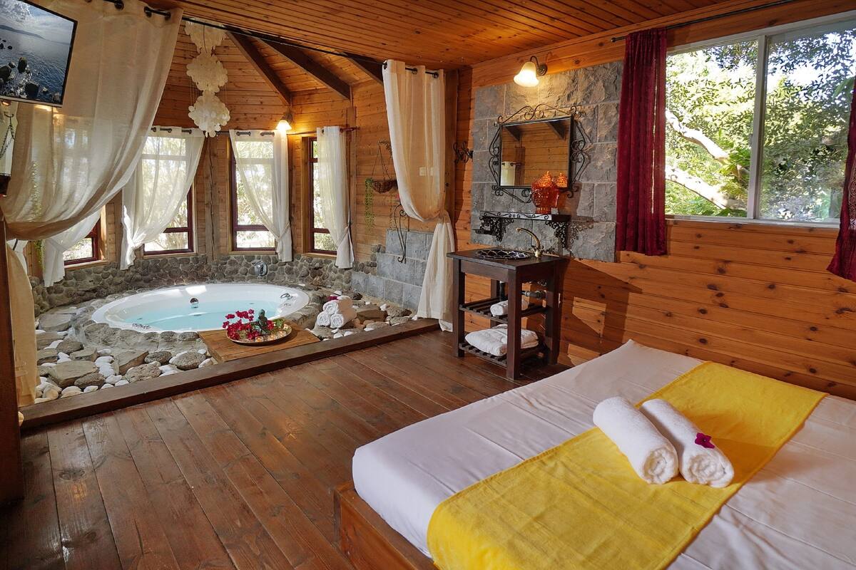 A wooden cabin with a stone wall, a wooden bed with a yellow blanket, a bathroom with a stone sink, and a hot tub surrounded by rocks.