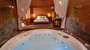 Una cabaña de madera con un dormitorio y un jacuzzi. La habitación tiene paredes de madera, una cama con almohadas de colores y una mesita de noche con una lámpara.