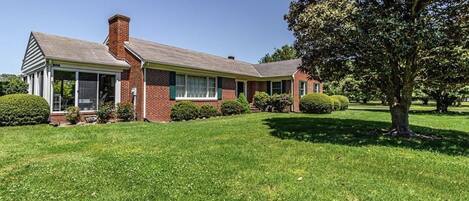 A two-story brick house with a covered porch, a well-maintained lawn, and a large tree in the front yard.