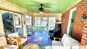 A sunlit room with a ceiling fan, a white sofa, two wooden chairs, a wicker ottoman, and a colorful rug. - Secluded, Waterfront Retreat: Fire pit, Private Dock, Game Room, Kayaks! (Reedville)