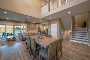 A spacious dining area with a long table and woven chairs, a kitchen in the background, and a staircase leading to a balcony. - Beach Bums Home with Pool, Steps to the Beach (Cape Charles)