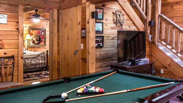 A pool table in a wooden cabin with a staircase and a television in the background. - Mountain Music 19 (Sevierville)