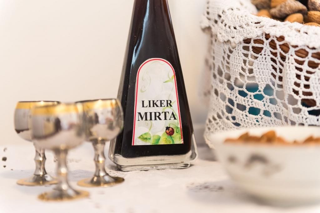 A bottle of Liker Mirta wine with a label featuring a ladybug and green leaves, placed next to a silver goblet and a crocheted basket filled with nuts.