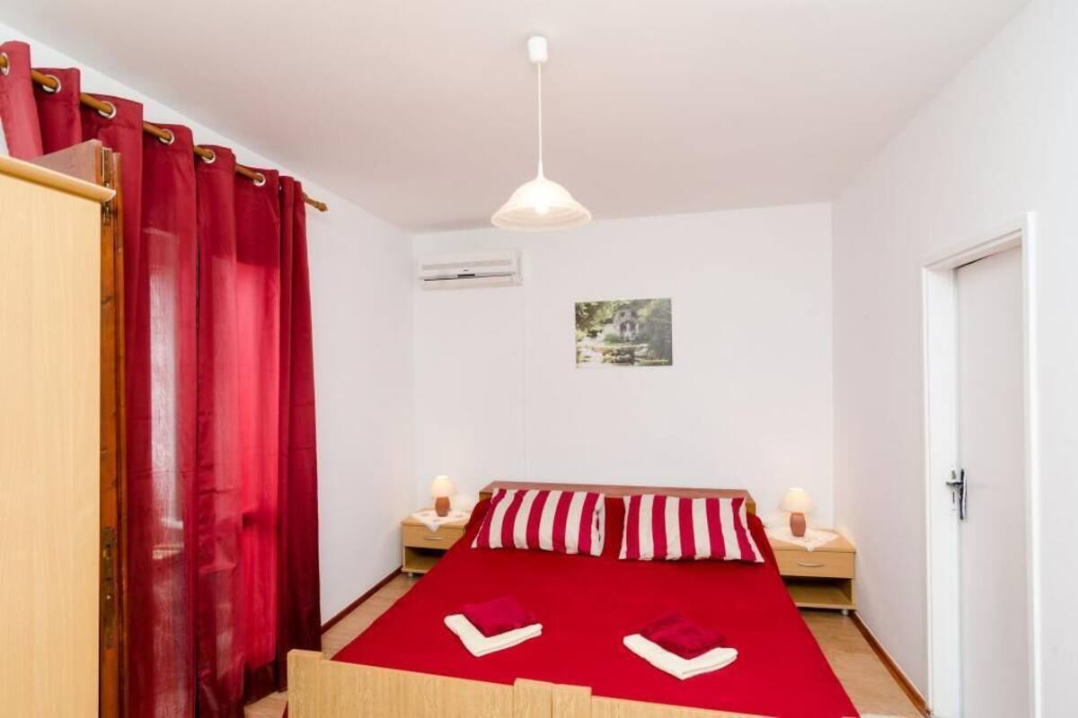A bedroom with a bed, red curtains, a white ceiling light, a small bedside table with a lamp, and a wall-mounted air conditioner.