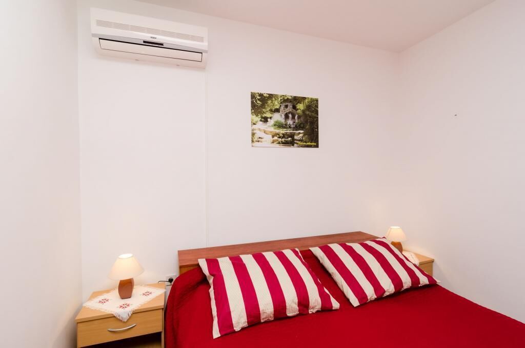 A bedroom with a single bed, a nightstand with a lamp, an air conditioner, and a wall-mounted picture.