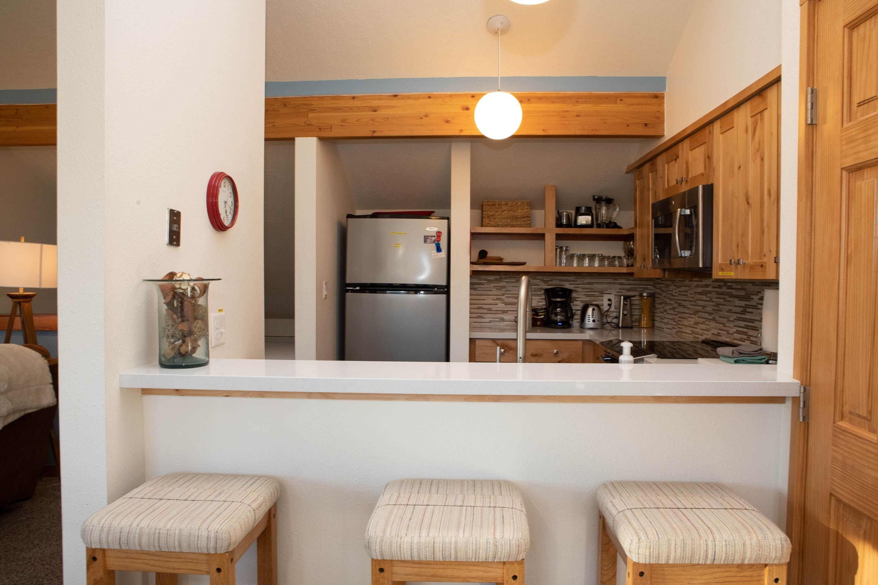 A compact kitchen with wooden cabinets, a stainless steel refrigerator, and a white countertop. There are three stools in front of the counter.
