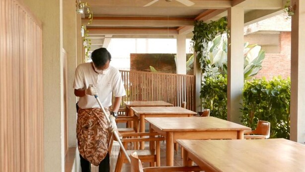 A clean, tiled corridor with wooden tables and chairs, a ceiling fan, and potted plants. - Ayom Java (Surakarta)