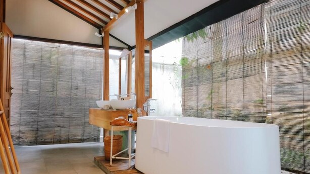 A modern bathroom with a freestanding bathtub, wooden beams, and a large window with blinds. - Ayom Java (Surakarta)