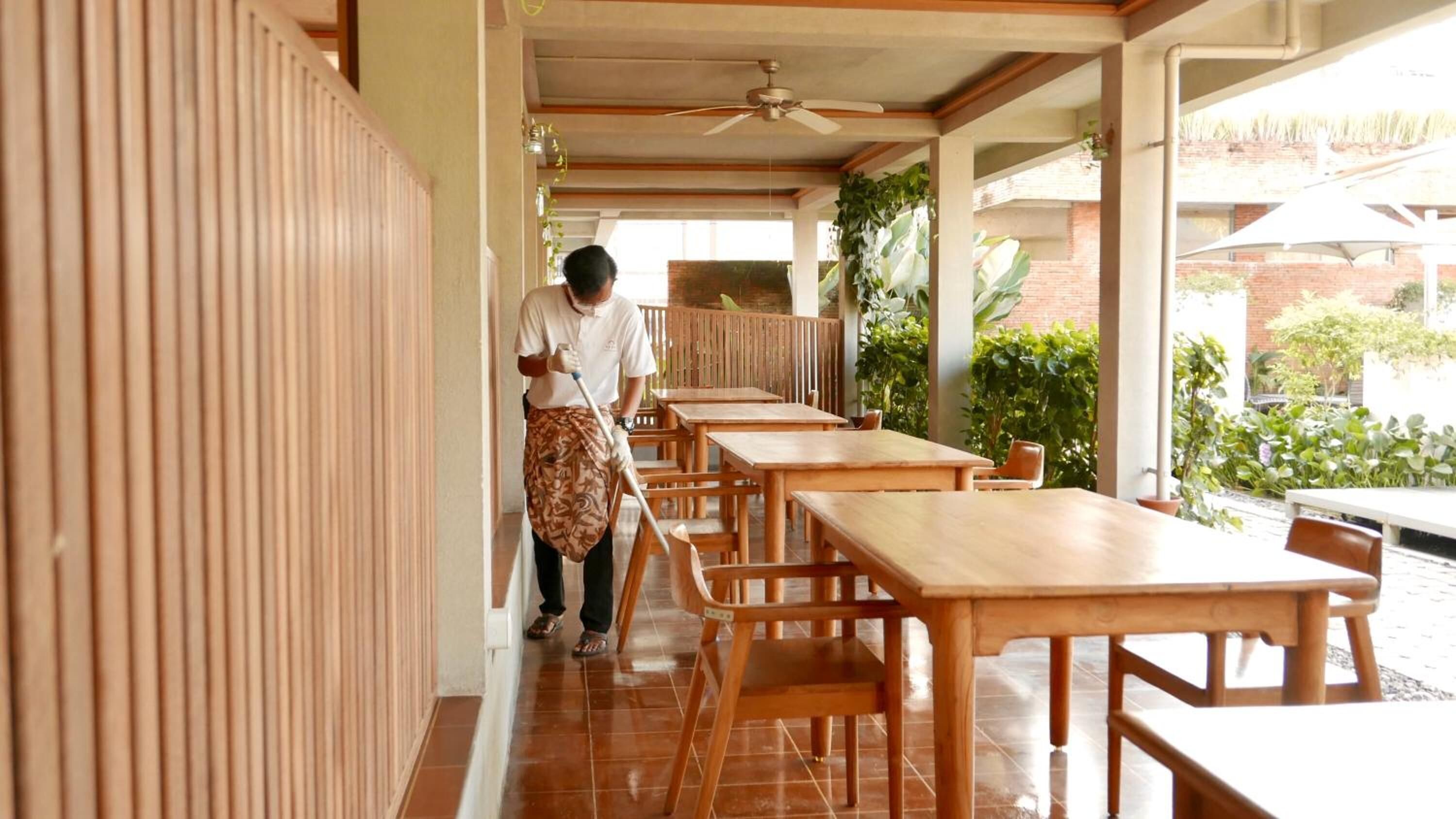 A clean, well-maintained corridor with wooden tables and chairs, a ceiling fan, and potted plants.