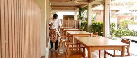 A clean, well-maintained corridor with wooden tables and chairs, a ceiling fan, and potted plants.