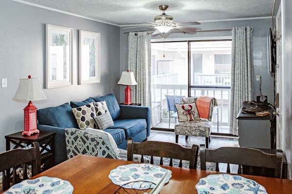 A living room with a blue sofa, a dining table with chairs, a ceiling fan, and a balcony with a view.