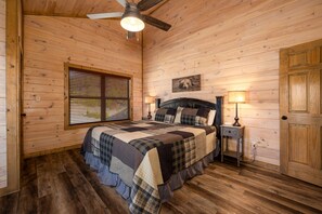 A wooden-paneled bedroom with a bed, a window with blinds, a ceiling fan, a bedside table with a lamp, and a door. - Stunning retreat: resort pool, theater, games, and mountain views for groups (Pigeon Forge)