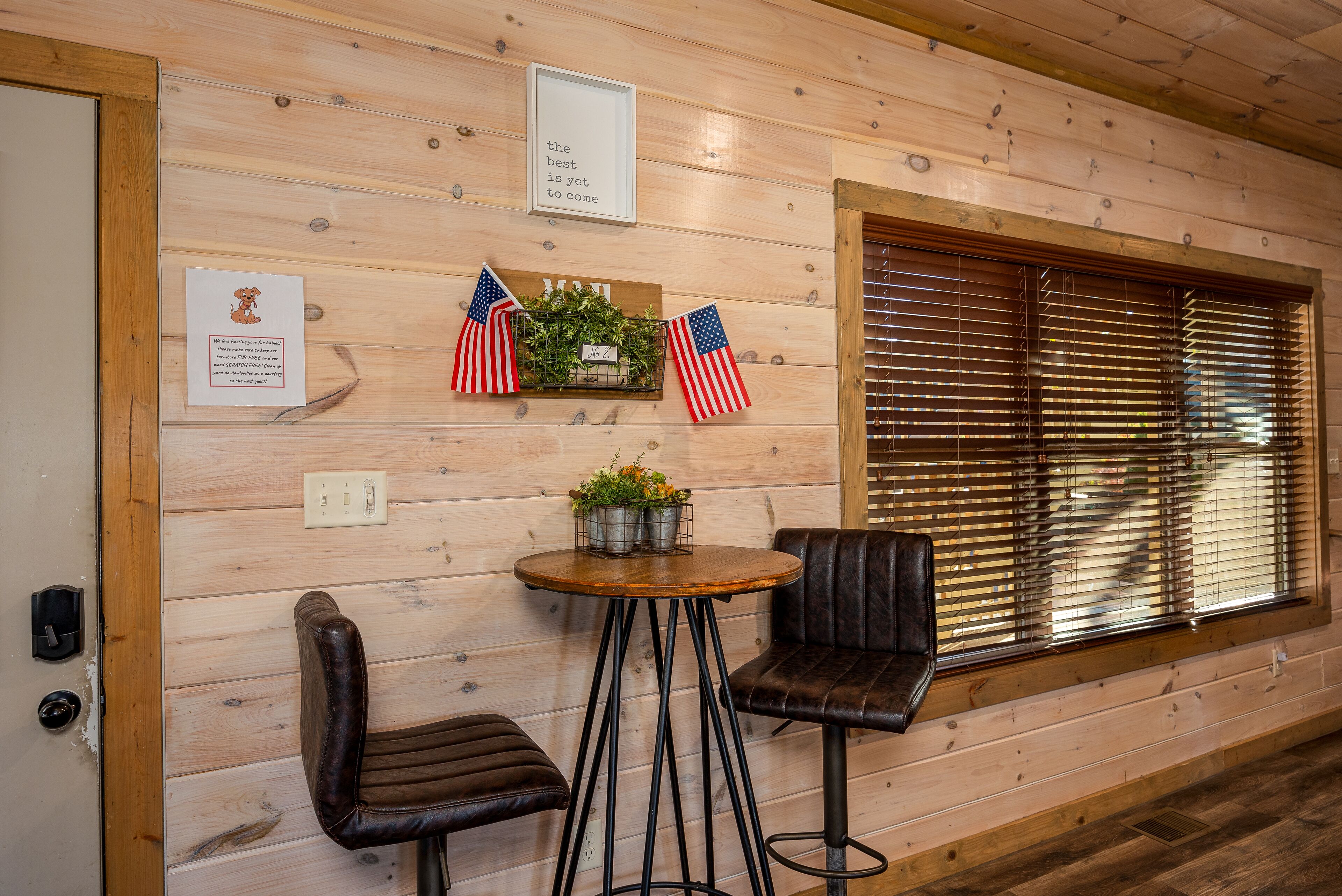 Interior de madera con ventana en marco de madera, dos sillones de cuero, una mesita redonda con una planta y un cartel con banderas estadounidenses.