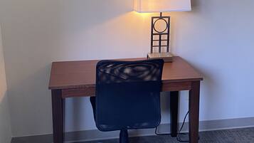 Et skrivebord med stol, lampe og et billede i ramme på væggen.