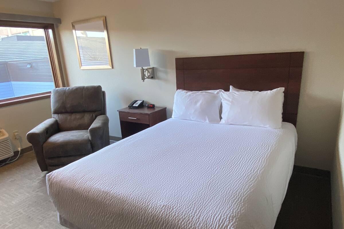 A hotel room with a large bed, a chair, a nightstand with a phone, and a window with blinds.