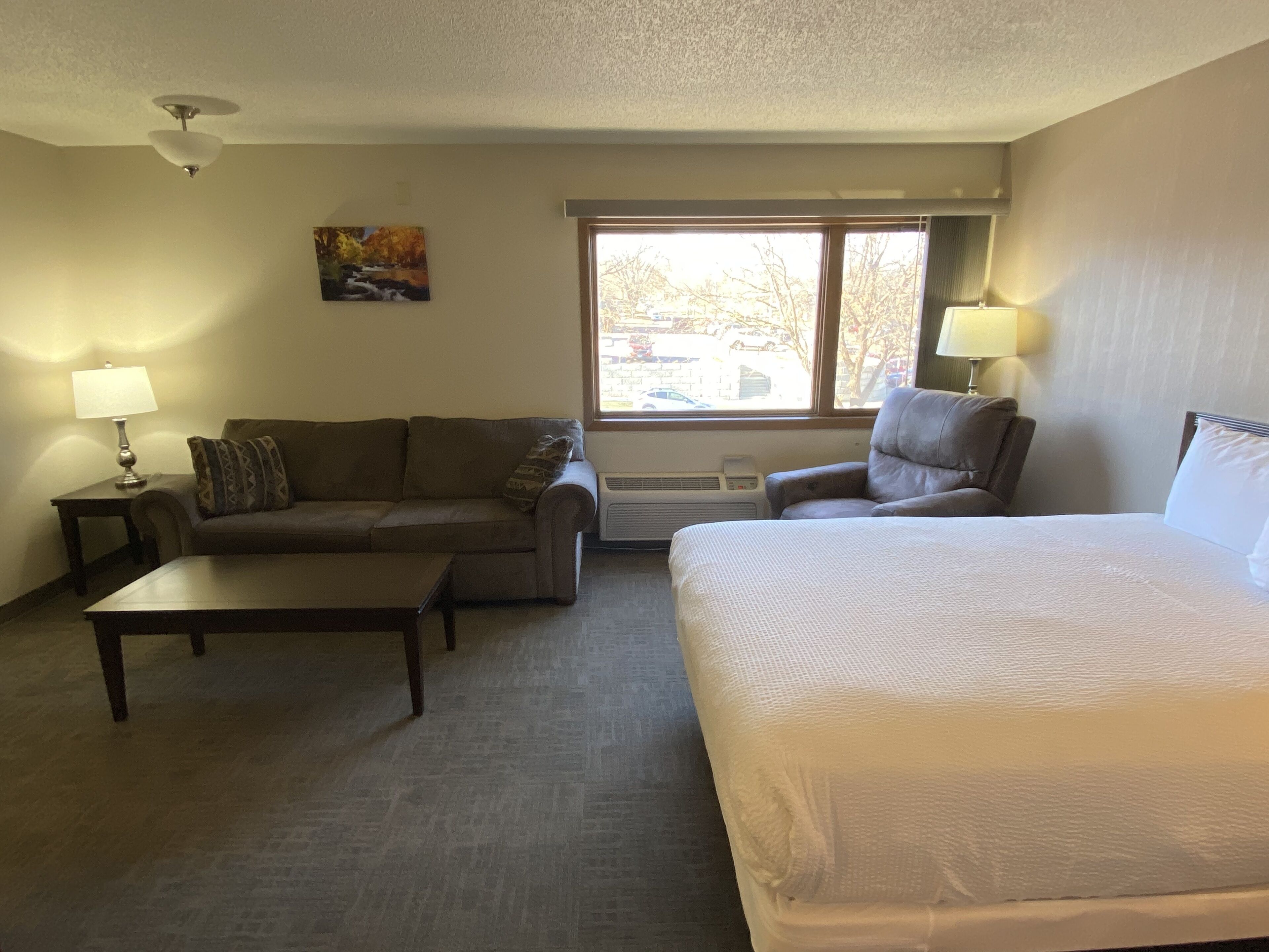 A hotel room with a bed, a sofa, a chair, a coffee table, and a window with a view of the outside.