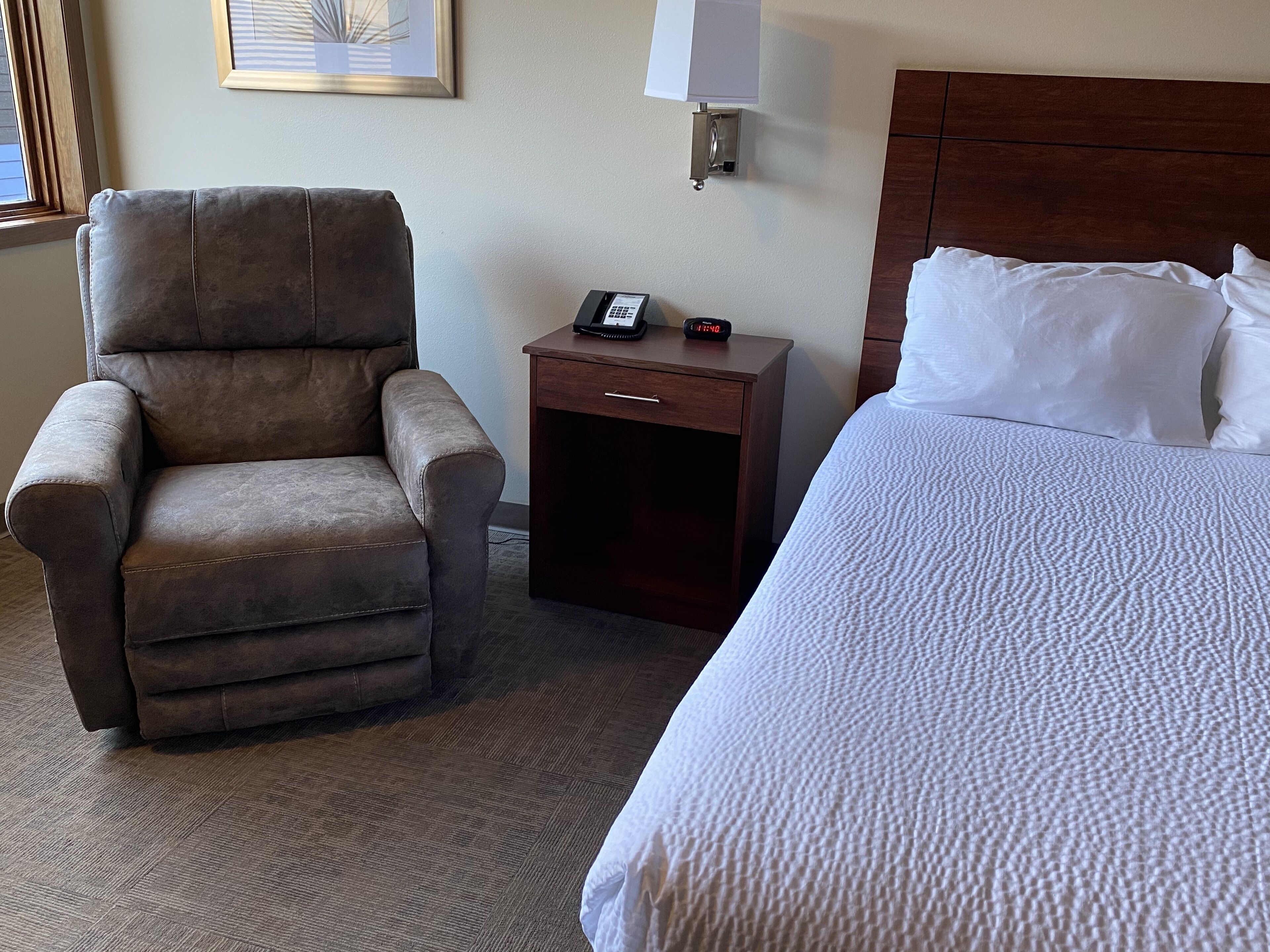 A hotel room with a large bed, a chair, a nightstand with a lamp, and a framed picture on the wall.