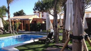 A pool area with lounge chairs and umbrellas, surrounded by palm trees and a building with a tiled roof. - House 1 suite Ragel coast (Palmar, El)
