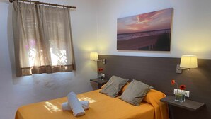 Et hotelværelse med en seng, to sengeborde med lamper, et sengebord med en vase med blomster, et vindue med gardiner og et maleri af en solnedgang på væggen.