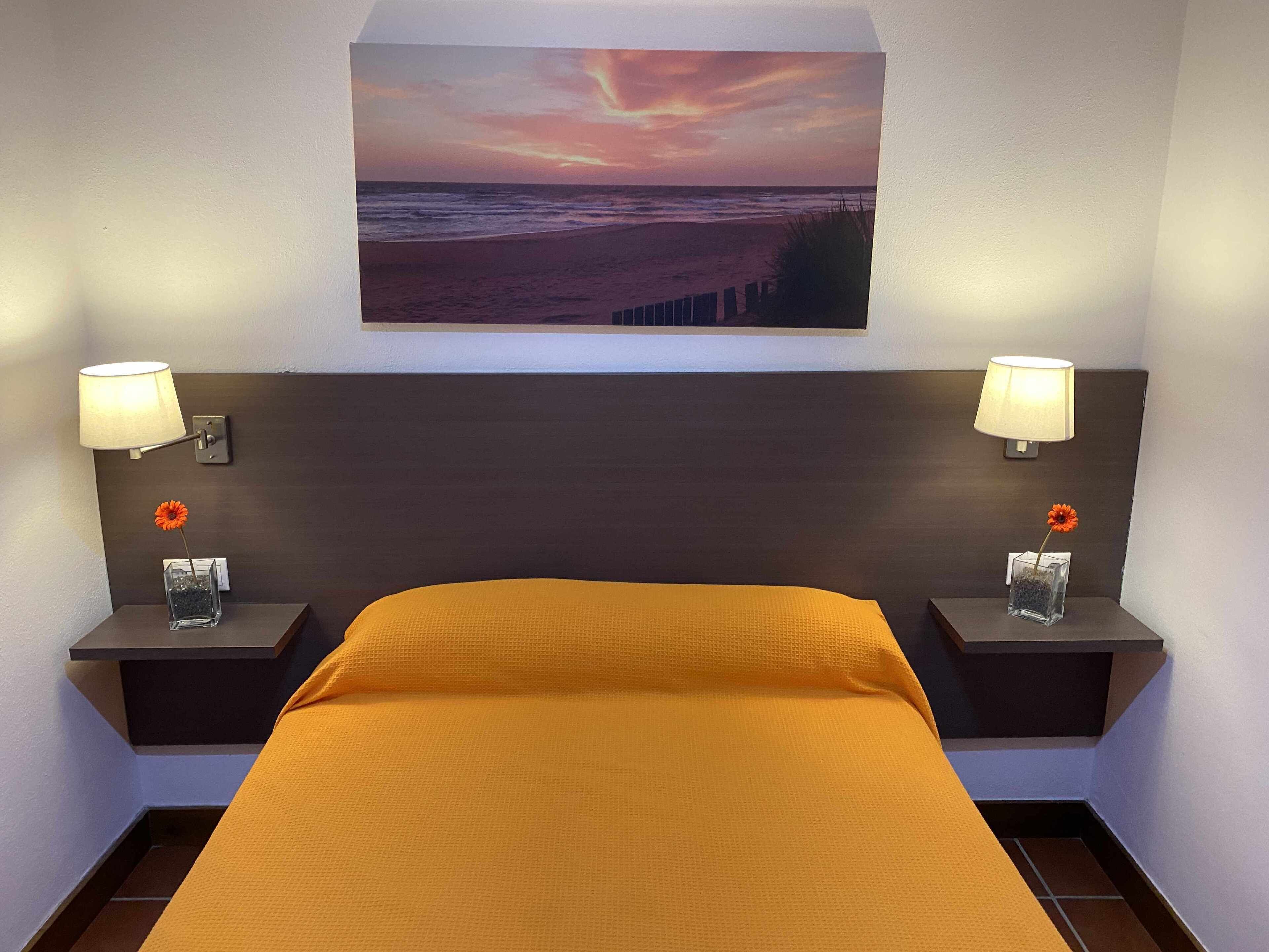 Una habitación de hotel con una cama, dos mesitas de noche con lámparas, un cuadro enmarcado con una puesta de sol sobre el océano y un pequeño florero con una flor en cada mesa.