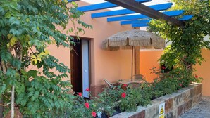 A covered outdoor seating area with a thatched umbrella, a stone planter with red flowers, and a blue pergola. - Apartamento con Doble Entrada y Piscina Compartida en el Palmar (Palmar, El)