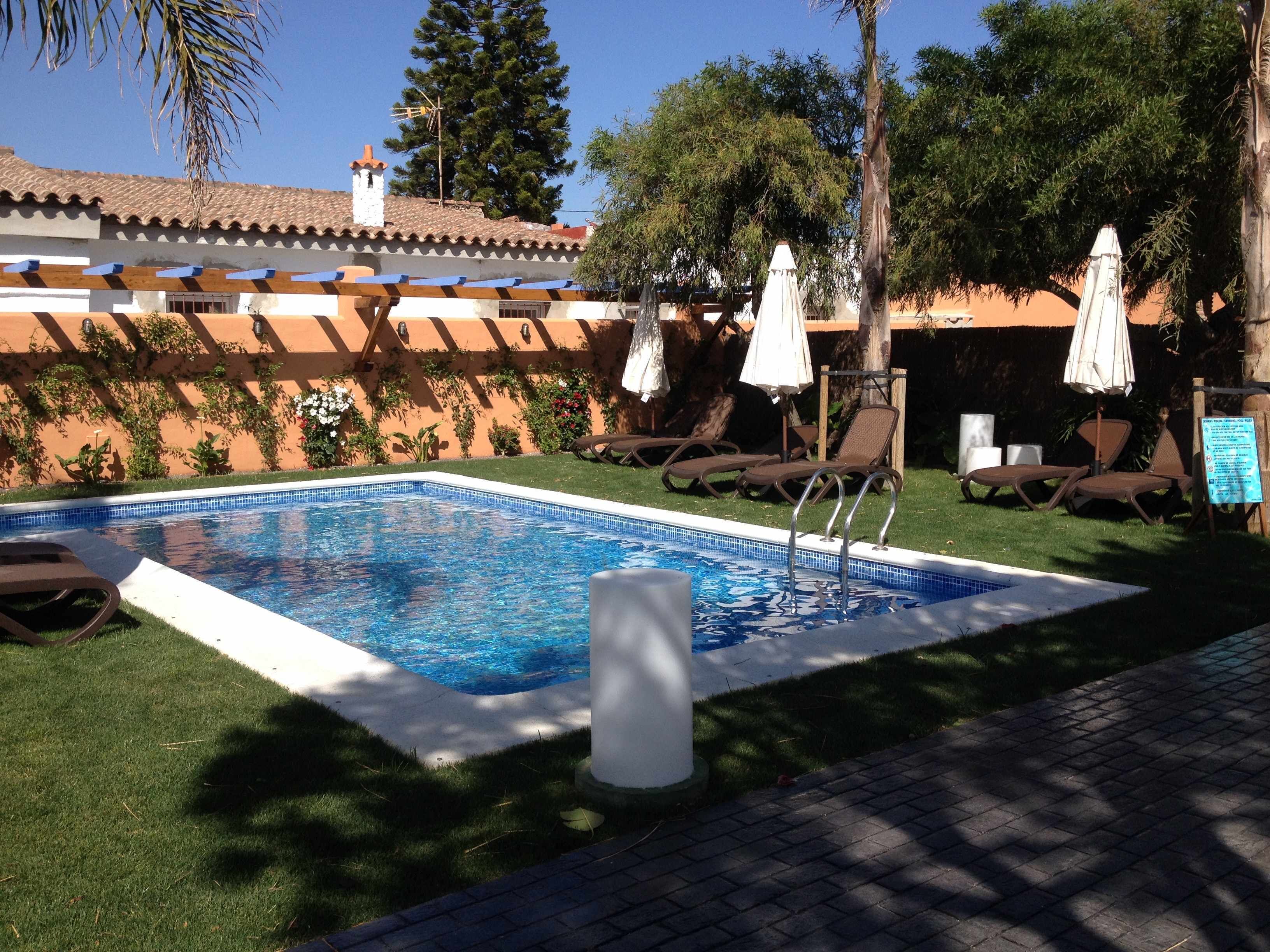 Une piscine entourée de verdure, dotée de chaises longues et de parasols, et surmontée d’une structure avec un toit en tuiles.
