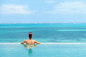 Una persona relajándose en una piscina infinita transparente, con vistas al océano y a la costa lejana.