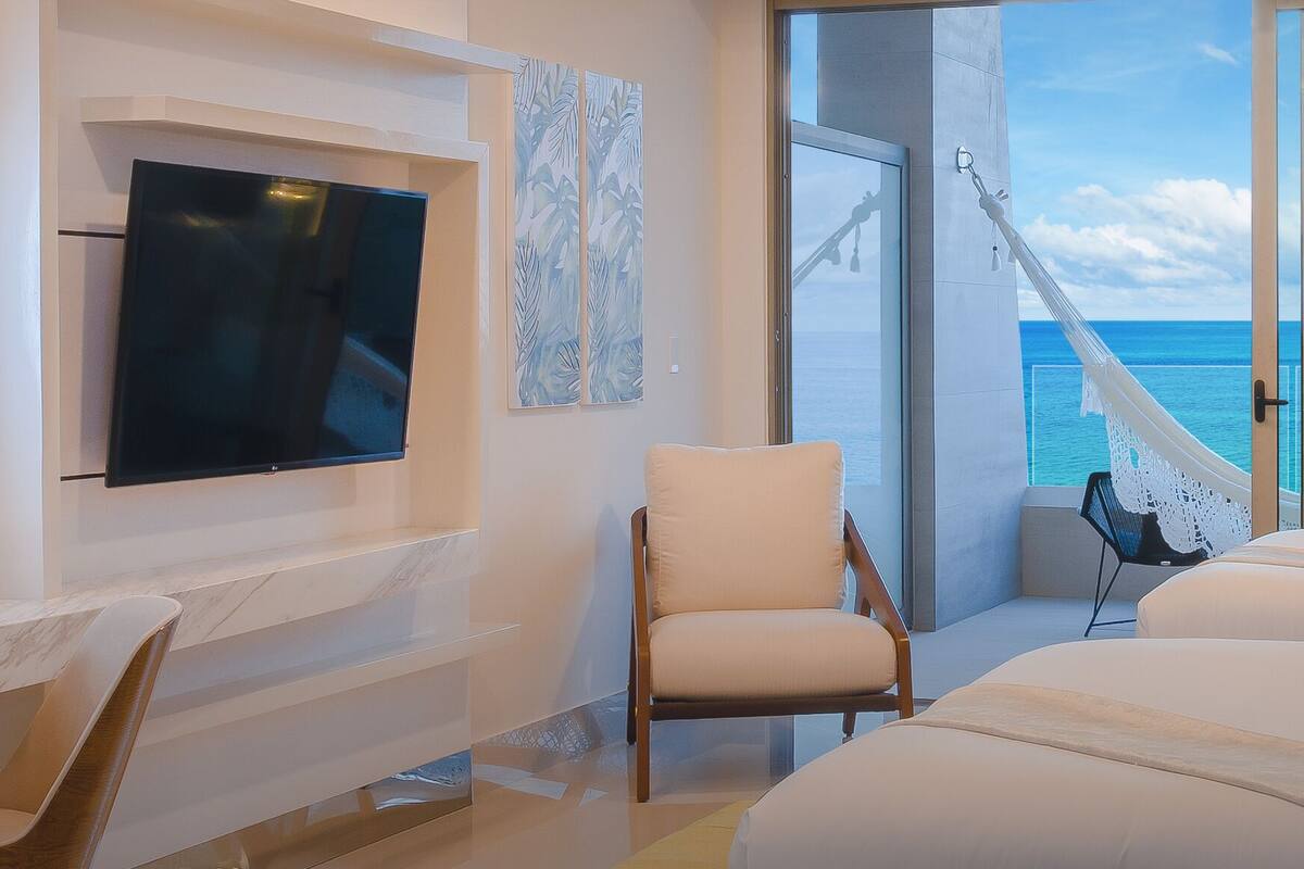 Habitación de hotel con televisor grande, una silla, cama con almohadas, ventana con vista al mar y una hamaca.