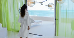 A room with a view of the sea, a white bed, and a green curtain.