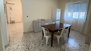 Un coin repas avec une table en bois et des chaises blanches, un réfrigérateur, un petit meuble et une fenêtre avec des stores.