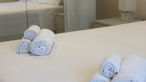 Un lit bien fait, avec des serviettes blanches pliées en rouleau et une table de chevet sur laquelle se trouve une lampe.