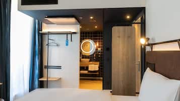 A modern hotel room with a large bed, a wooden wardrobe, a built-in circular mirror, and a small bathroom with a sink and towel rack.
