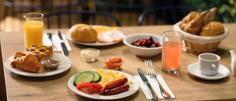 Una mesa de desayuno con croissants, salchichas, tomates, pepinos y un cuenco de bayas.