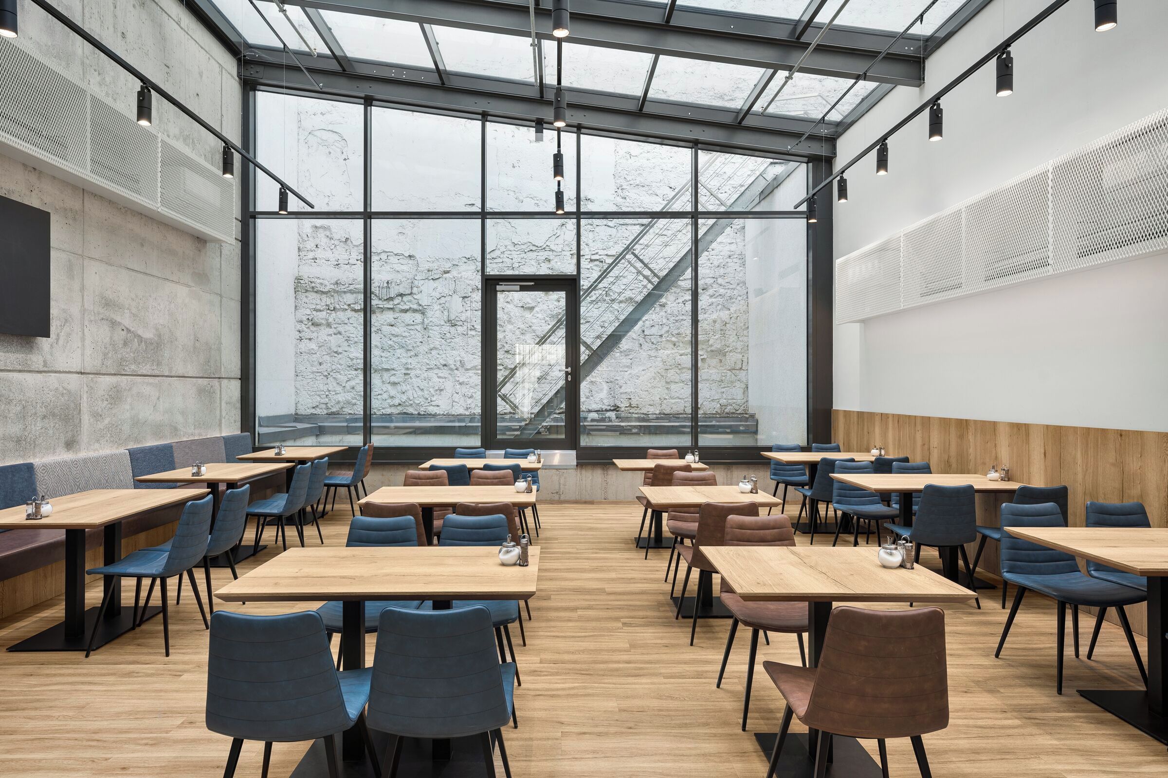Un vaste espace de restauration intérieur, doté de hauts plafonds et de grandes fenêtres, et présentant un design moderne avec des tables en bois et des chaises rembourrées.
