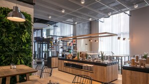 Een modern café met houten tafels, een centraal eiland met een koffiebar en een groene wand met planten.
