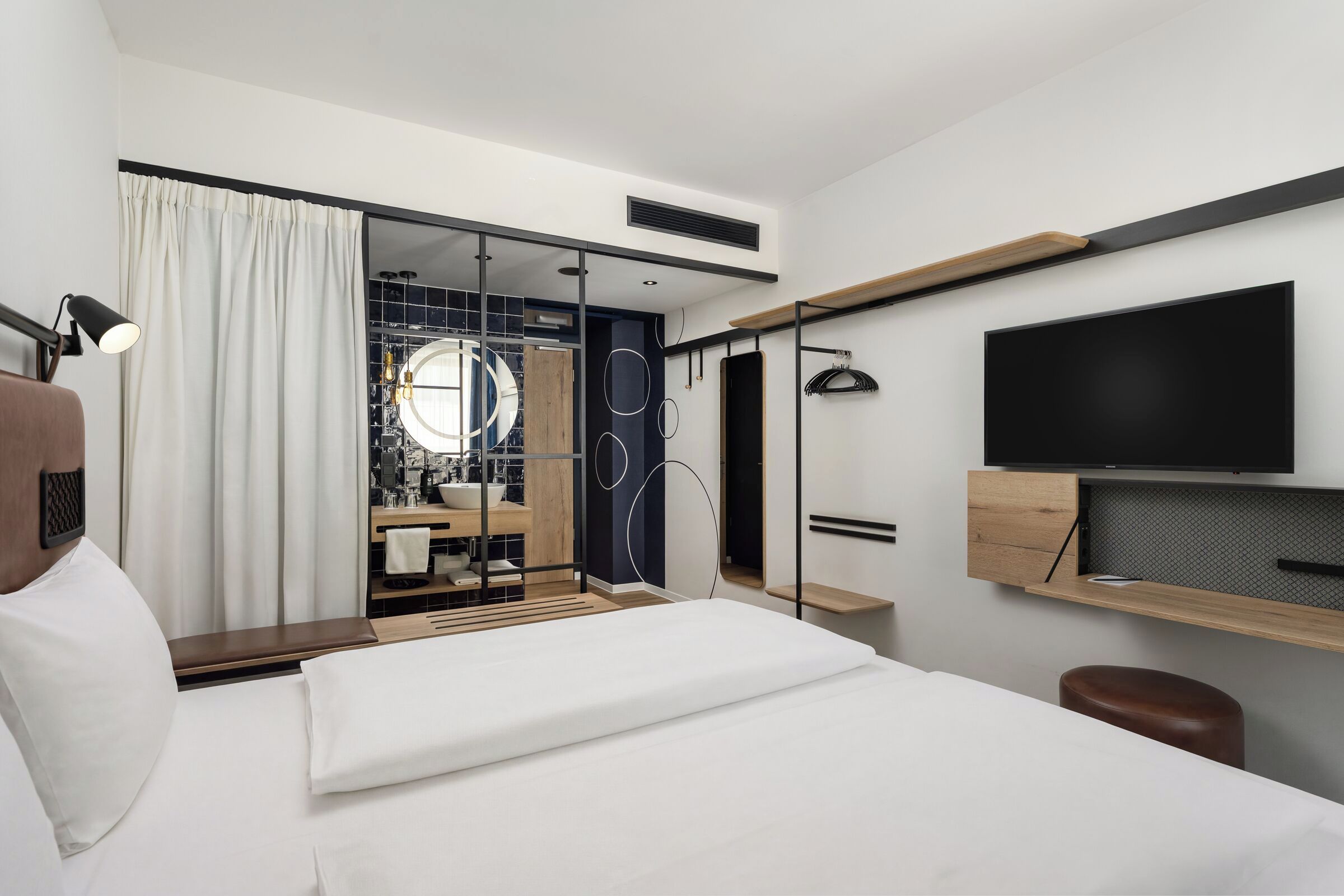 A modern hotel room with a bed, a TV, a desk, and a bathroom visible through a glass door.