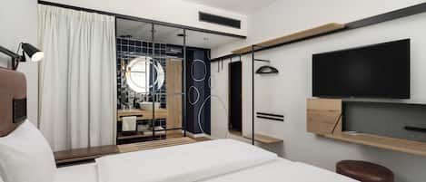 Une chambre d’hôtel moderne équipée d’un lit, d’une télévision, d’un bureau et d’une salle de bain visible à travers une porte en verre.
