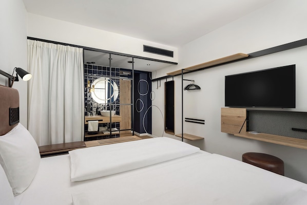 Habitación de hotel moderna con cama, televisor, escritorio y baño visible a través de una puerta de cristal.