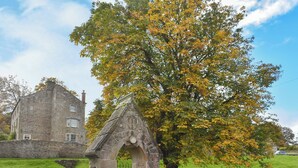 A stone structure with an archway, a stone building, and a large tree with yellow leaves in a grassy area.