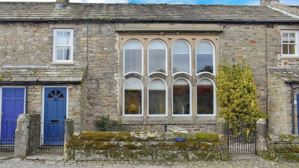 A stone building with a large arched window, a blue door, and a small garden area with a moss-covered stone wall.