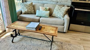 Una sala de estar con un sofá, una mesa de centro con un libro y un control remoto, una cocina con un horno y una ventana con cortinas.