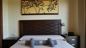 Een netjes opgemaakt bed met opgerolde handdoeken, een donker houten hoofdbord, een aan de muur hangend kunstwerk, een nachtkastje met een lamp en een nachttafeltje.