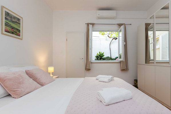 A bedroom with a bed, pillows, a window with curtains, a bedside lamp, and a wall-mounted air conditioner. - Rooms Batina (Dubrovnik)