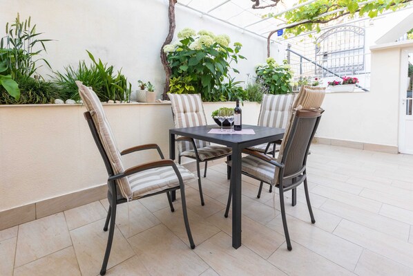 Un patio al aire libre con una mesa y sillas, rodeado de plantas en macetas y una pared blanca.