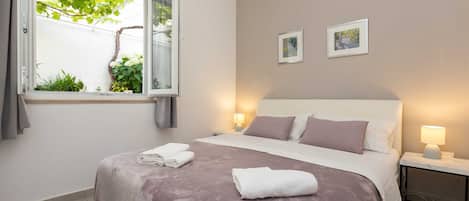 A bedroom with a bed, bedside lamps, a window with a view of greenery, and two framed pictures on the wall.