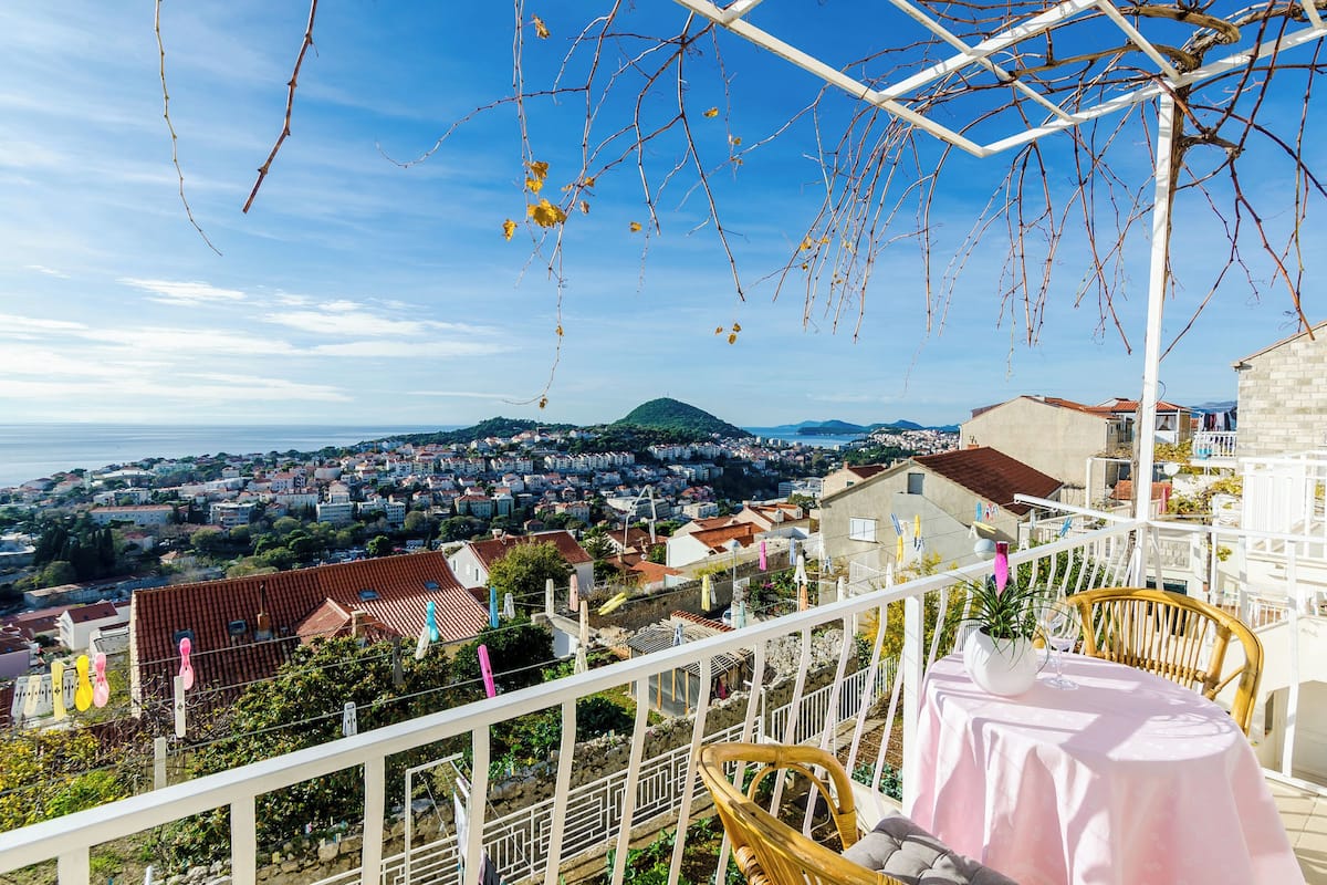 A balcony with a table and chairs, overlooking a coastal town and a mountain.