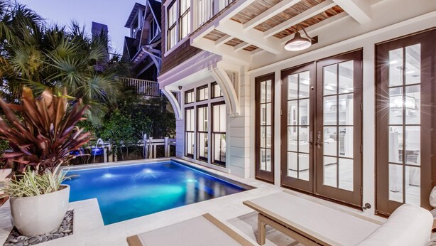 Un área de piscina moderna con una piscina azul cristalina, rodeada de mobiliario exterior blanco y grandes puertas de vidrio que dan a un interior bien iluminado.