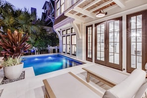 Un área de piscina moderna con una piscina azul cristalina, rodeada de mobiliario exterior blanco y grandes puertas de vidrio que dan a un interior bien iluminado.